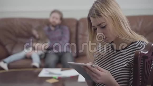美丽的金发女人坐在丈夫和小女孩模糊的身影面前手里拿着平板电脑的画像视频的预览图
