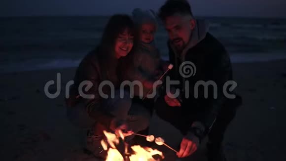 妈妈爸爸穿着暖和的衣服正在用木棍和他们的小宝宝烤棉花糖海滩上的篝火视频的预览图