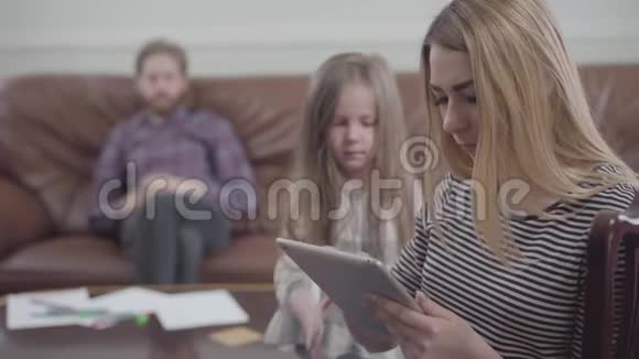 一幅漂亮的金发女人坐着的肖像手里拿着平板电脑小女孩想引起妈妈的注意视频的预览图