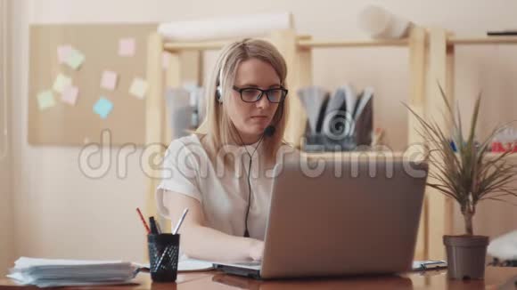 戴着眼镜和一件白色上衣的白种人可爱的女孩为笔记本电脑工作耳机正在交流视频的预览图