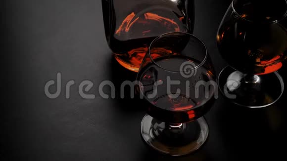 豪华白兰地带有金色干邑的圆形瓶子和两个玻璃杯在黑色背景下的黑色桌子上旋转玻璃眩光视频的预览图