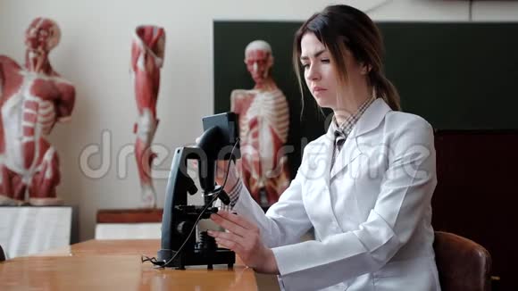 一个女人坐在显微镜后面在人体模型的背景下研究人的结构视频的预览图