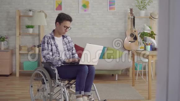 一位戴着时尚眼镜的亚洲青年男子坐在轮椅上带着笔记本电脑在房子的客厅里视频的预览图