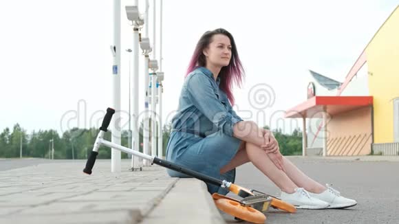 迷人的年轻女人染了头发穿着牛仔裙和白色运动鞋坐在路边休息视频的预览图