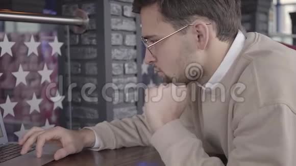 戴眼镜的年轻帅哥坐在餐桌上的餐馆里用笔记本电脑打字视频的预览图