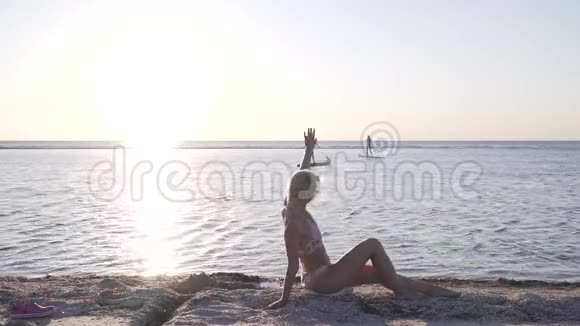 海滩上一位穿比基尼的女士正在海上冲浪板上向两位女士挥手视频的预览图
