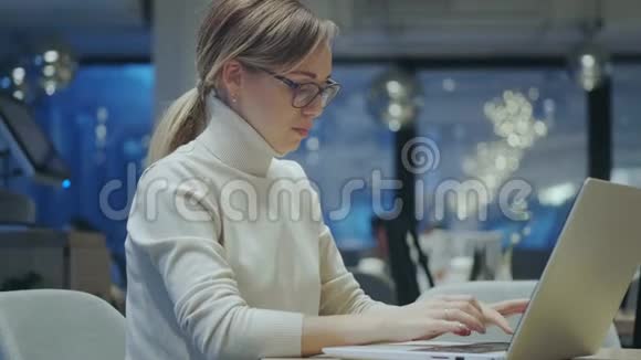 戴着眼镜的漂亮女人坐在一家晚上的咖啡馆里用笔记本电脑工作视频的预览图