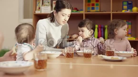幼儿园老师和孩子们一起坐在桌子旁用勺子喂那个黑发小男孩另一个视频的预览图