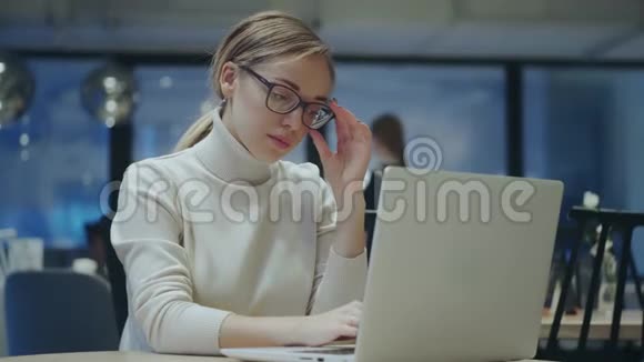 戴眼镜的年轻女子坐在一家晚上的咖啡馆里在笔记本电脑上工作视频的预览图