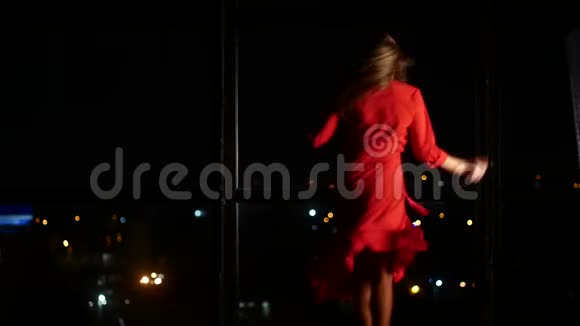 一个穿着红色优雅连衣裙的女孩正在一扇大窗户和一个夜晚的城市的背景下跳舞独自在家和视频的预览图