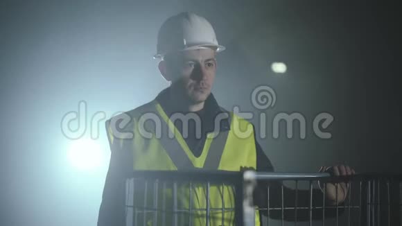 肖像微笑的人穿着建筑工服和头盔站在黑色背景前聚光灯下视频的预览图