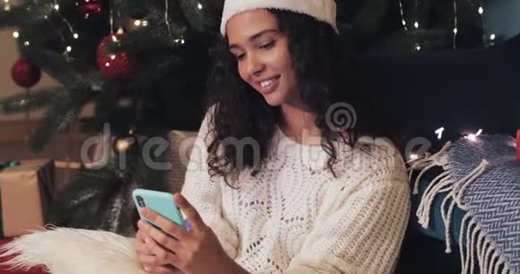 在节日装饰的家庭背景下一位戴着桑塔斯帽子的年轻女子坐在圣诞树下视频的预览图