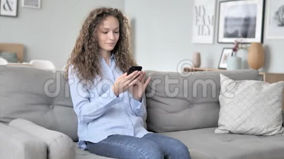 年轻的卷发女士在沙发上放松时使用智能手机视频的预览图