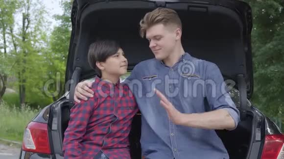 年轻的父亲和他的儿子坐在户外的汽车后面拥抱男孩的男人人们互相看着视频的预览图