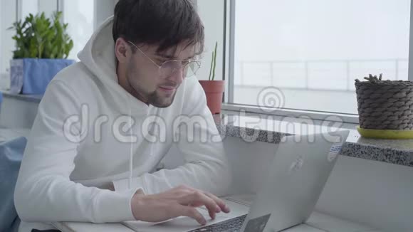 一个年轻时尚的英俊留胡子的人穿着一件轻便的运动衫坐在桌子旁用笔记本电脑打字视频的预览图