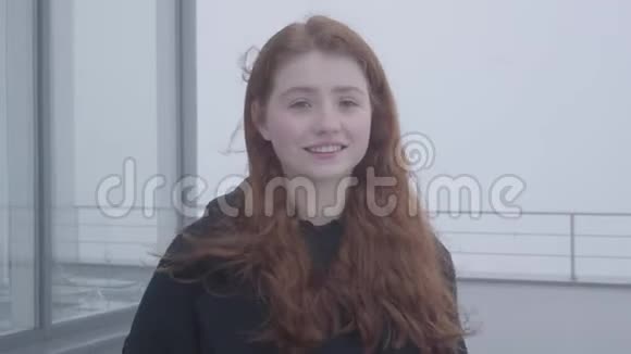 一个年轻可爱的女孩的肖像她穿着一件深色外套红头发站在屋顶或露台上在寒冷多风视频的预览图