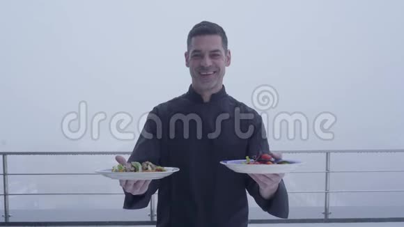 黑色制服的快乐成人职业厨师的肖像两个盘子在雨雾的背景下视频的预览图
