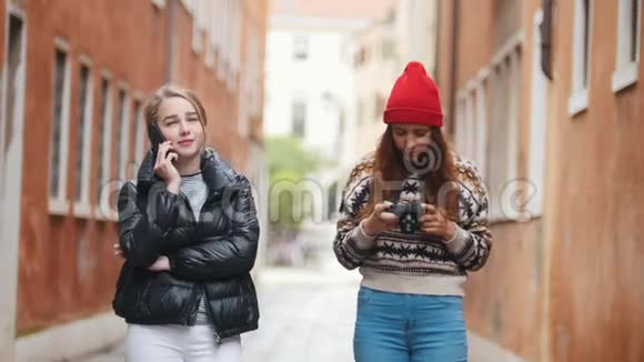 两个穿着温暖衣服的年轻女性旅行者走在狭窄的街道上一个女人在打电话另一个视频的预览图