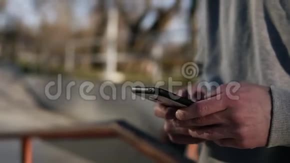 阳光明媚的一天年轻卷曲的头男滑板运动员在滑板公园里玩手机靠在栏杆上视频的预览图