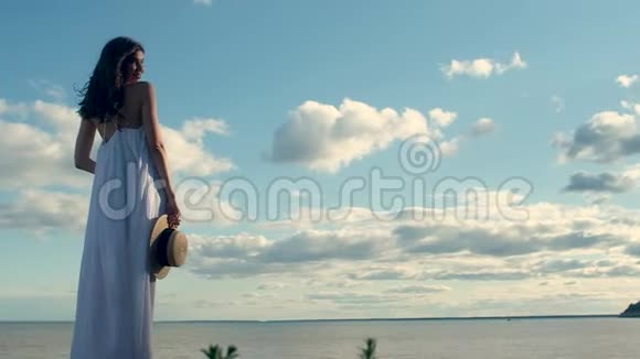 美丽的女人穿着白色长裙手里拿着帽子在大海的背景上视频的预览图