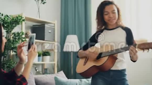 可爱的年轻女子业余吉他手正在弹吉他和唱歌而她的亚洲朋友正在录制视频视频的预览图