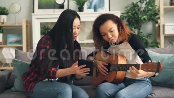 混合种族的朋友亚裔和非裔美国人正在学习使用平板电脑弹吉他女孩们看着屏幕视频的预览图
