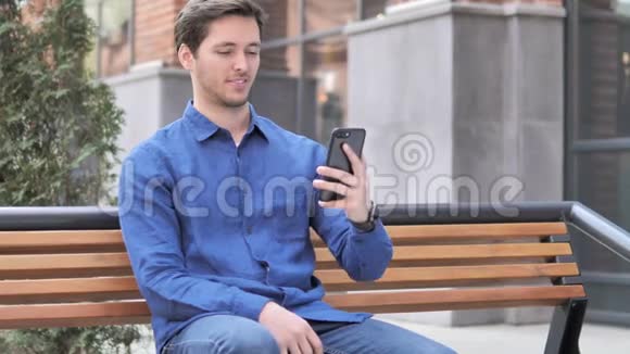 青年户外坐著的智能电话视频聊天视频的预览图