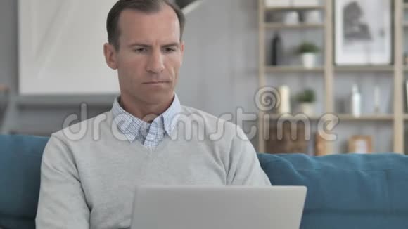 沮丧的老人在坐在沙发上用笔记本电脑面对损失视频的预览图