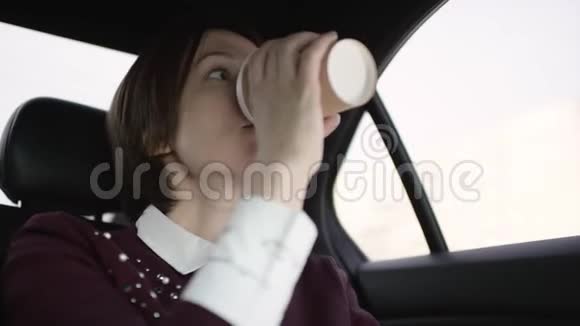 穿着白色上衣和栗色毛衣的棕色头发的漂亮女人在车里堵车喝咖啡视频的预览图