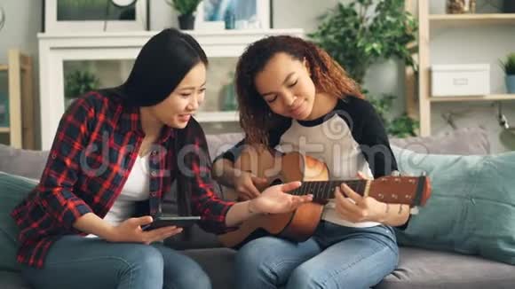 亚洲女人正在教她的非裔美国朋友在家里闲暇时间弹吉他一个女孩抱着视频的预览图
