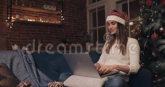 微笑迷人的女人戴着圣诞帽坐在家背景的圣诞树附近利用她的笔记本聊天视频的预览图