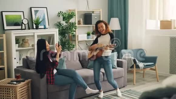 可爱的非裔美国妇女正在家里弹吉他而她的亚洲朋友正在用智能手机和手机录制视频视频的预览图
