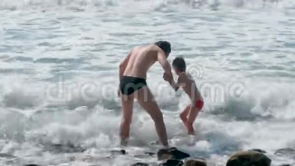 爸爸和儿子在石滩上的暴风雨中在海浪中游泳后景视频的预览图
