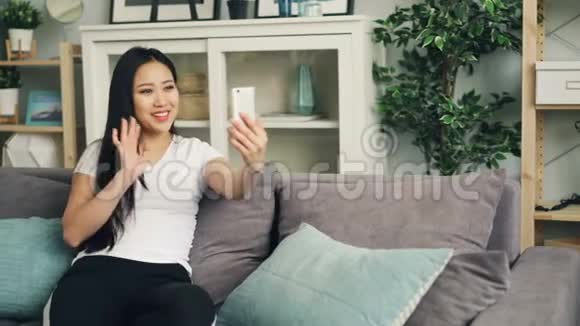 快乐的亚洲年轻女士在家中独自坐在客厅的沙发上用现代智能手机进行在线视频通话视频的预览图