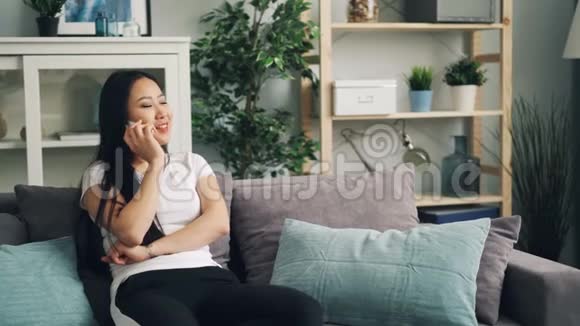 漂亮的亚洲女孩正坐在漂亮的现代公寓的沙发上用手机笑着说话视频的预览图
