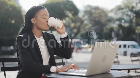 微笑的非裔美国女商人坐在夏日露台的咖啡馆里喝着咖啡在笔记本电脑上工作视频的预览图