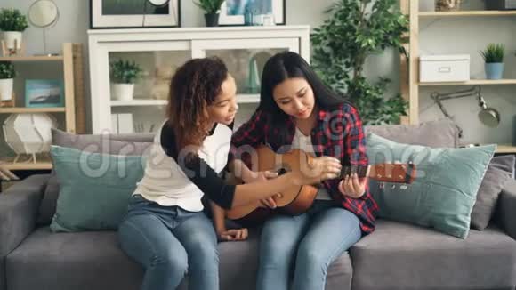 美丽的亚洲女孩穿着休闲服装正在学习弹吉他而她的非裔美国朋友年轻女士则是视频的预览图