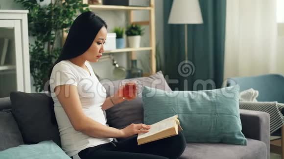 漂亮的亚洲女人正在看书享受现代文学坐在沙发上在家喝茶放松视频的预览图