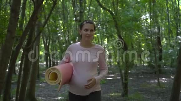 做瑜伽的孕妇一个女孩走在公园里手里拿着地毯另一只手放在肚子上视频的预览图