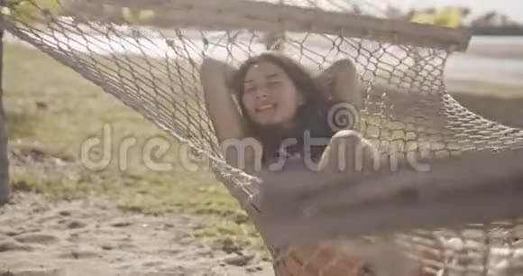 在海滩上一个漂亮开朗的年轻女孩躺在吊床上穿着黑色性感比基尼穿着宽大的比基尼微笑视频的预览图