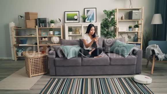 年轻的亚洲女性正在使用智能手机在沙发上休息而机器人吸尘器则在用吸尘器打扫公寓视频的预览图