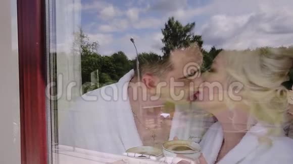 年轻人和女孩在窗户附近的玻璃后面接吻他们都穿着白色晚礼服手中一杯咖啡视频的预览图