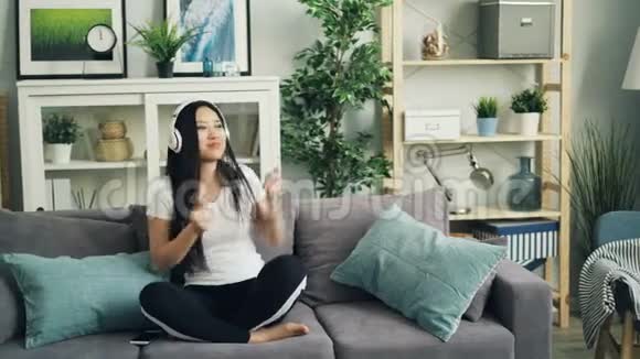 穿休闲服装的快乐年轻女士坐在家里的沙发上听着音乐戴着耳机跳舞唱歌视频的预览图