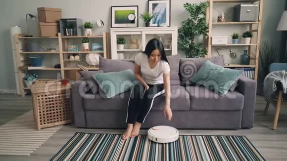 美丽的亚洲女孩打开机器人吸尘器然后坐在沙发上使用智能手机享受方便视频的预览图