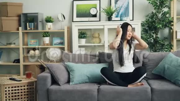 美丽的亚洲女孩正通过耳机听音乐跳舞手和身体在家坐在沙发上视频的预览图
