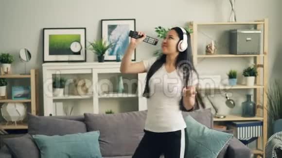 可爱的亚洲女孩戴着无线耳机在遥控器里唱歌在客厅里跳舞在舒适的公寓里放松和放松视频的预览图
