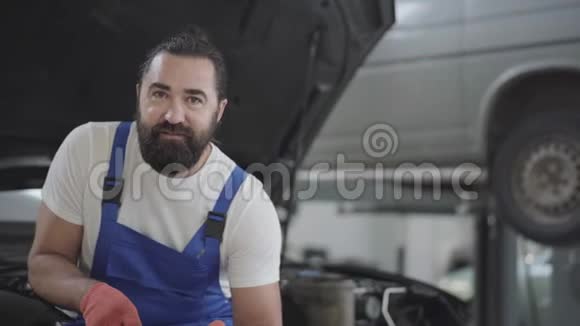 在一家汽车修理店里有着迷人的胡须的机械师摆姿势扳手坐在一辆破车的引擎盖上视频的预览图