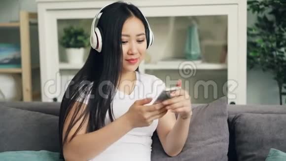 可爱的亚洲年轻女性正在听耳机里的音乐触摸着家中沙发上的智能手机屏幕视频的预览图