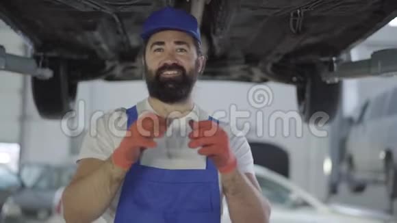 成年机械师拿出手套站在被提起来的车下微笑着穿着制服装汽车的胡子男视频的预览图