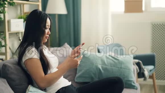 有魅力的亚洲女士使用智能手机用银行卡在家里坐在沙发上购物视频的预览图